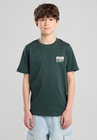 T-paita tummanvihreää puuvillaa, lyhyet hihat, ribbikuvio, pyöreä kaula-aukko ja valkoinen "Hyvä Elämä" -grafiikka, jossa on kukkakuvio.