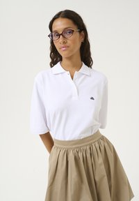Cămașă polo albă cu mâneci scurte și un logo brodat de culoare neagră, asortată cu o fustă plisată bej, care prezintă o textură netedă a țesăturii.