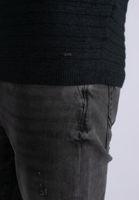 Pull en texture sombre avec des côtes horizontales et une étiquette noire en bas, associé à un jean en denim gris ajusté avec des détails légèrement déchirés.
