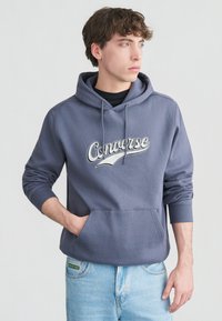 Νεαρός άνδρας που φοράει μπλε φούτερ Converse με το χέρι στην εμπρόσθια τσέπη, κοιτάζοντας δεξιά, συνδυασμένο με ανοιχτόχρωμο τζιν μπροστά σε απλό φόντο.