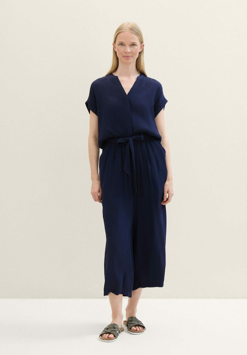 Salopette bleu marine à manches courtes, décolleté en V et ceinture nouée à la taille, avec des jambes larges et un tissu texturé. Portée avec des sandales olive.