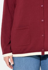 Hand som bär en vinröd knäppt cardigan med framficka över ljusblå jeans mot vit bakgrund.