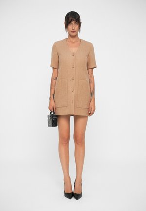 Abito corto in misto lana color tan, con maniche corte, chiusura con bottoni, due tasche e una silhouette aderente. Indossato con tacchi neri e una borsa piccola.
