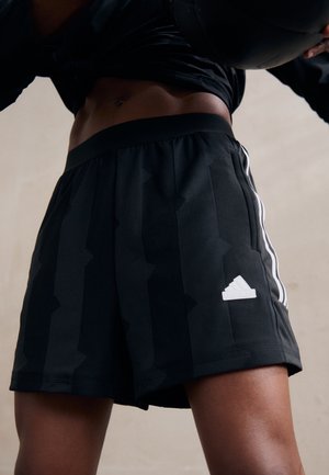 Osoba nosząca czarne spodenki Adidas z pionowymi paskami trzymająca czarną piłkę nad głową, widoczna od talii do połowy uda.