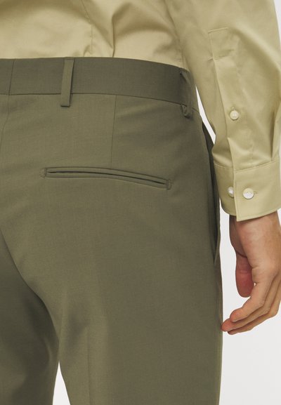 Olivgrüne Hose mit glatter Textur und Slim-Fit. Mit einer Reißverschlusstasche auf der Rückseite, kombiniert mit einem hellbeigen Hemd.