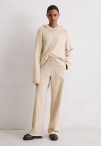Haut à capuche beige en tricot avec une capuche et des manches larges, associé à un pantalon en velours côtelé beige clair à jambe large, sur fond neutre.