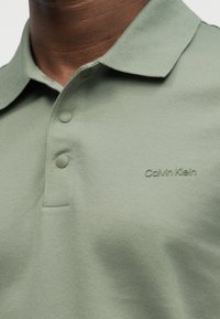 Άνδρας που φοράει πουκάμισο πόλο σε χρώμα φασκόμηλου με κουμπιά σκράπ και ανάγλυφο λογότυπο Calvin Klein στο στήθος.