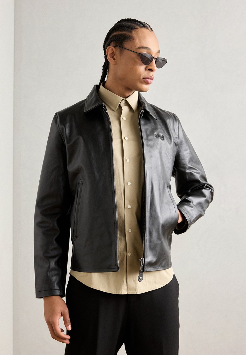 Sort læderjakke med lynlås, spidse krave og sidelommer, lagdelt over en beige skjorte med knapper. Glat tekstur, moderne pasform.