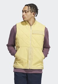 Muž s vrkočmi, ktorý má na sebe žltú prešívanú vestu cez fialovú košeľu s dlhým rukávom a tmavomodré nohavice, pozerá sa doľava s rukami v vreckách.