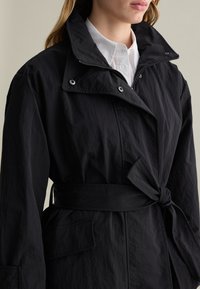 Abrigo negro de trench con un alto cuello, cintura ceñida y grandes bolsillos. Confeccionado con una tela texturizada que muestra las costuras y cierres a presión visibles.