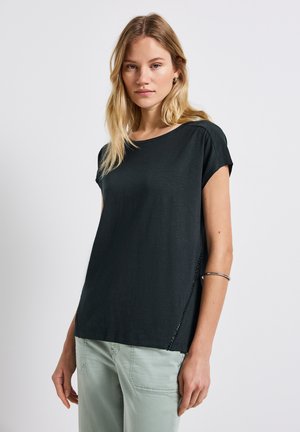 T-shirt nera a maniche corte con vestibilità rilassata, caratterizzata da spacchi laterali e una texture liscia. Indossata con pantaloni verde chiaro.