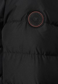 Czarna tkanina kurtki puchowej o pikowanej fakturze z okrągłą naszywką z logo na środku, obramowaną czerwonymi detalami.