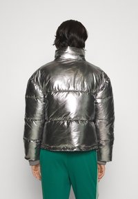 Stříbrná kovová prošívaná bunda s vysokým límcem a krabicovým střihem. Má prošívaný vzor a silnou výplň pro izolaci.