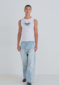 Hvit ermeløs tanktopp med et svart grafisk design på brystet, kombinert med lyseblå, løstsittende jeans og svarte støvler.