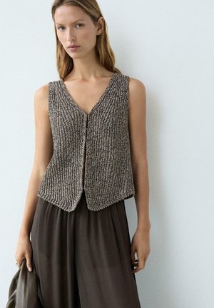 Donna che indossa un gilet senza maniche in maglia strutturata sopra pantaloni larghi scuri, in piedi contro una parete chiara e semplice, con una giacca in una mano.