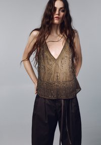 Gyöngyös olívaszínű ujjatlan felső mély V-nyakkivágással, bonyolult hímzéssel és laza szabással. Sötét, bő szárú nadrággal párosítva.