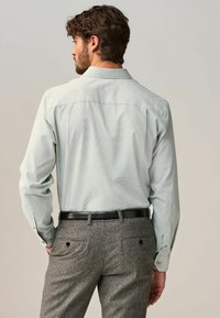 Hellgrünes gemustertes Hemd mit langen Ärmeln und Knopfmanschetten, kombiniert mit grauen, maßgeschneiderten Hosen und einem schwarzen Gürtel.