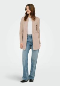Blazer long beige à texture lisse, ouvert à l'avant sans boutons, associé à un jean évasé bleu clair et des chaussures noires.