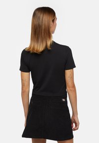 Camiseta de algodón negra de manga corta, combinada con una falda de pana negra que tiene un parche con el logo y bolsillos traseros, vista desde atrás.