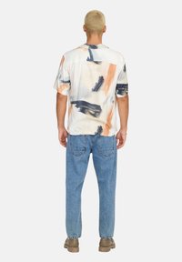 Camisa de manga corta en una tela suave y ligera que presenta un estampado de salpicaduras de pintura multicolores. Combinada con unos vaqueros sueltos de color azul y zapatos marrones.