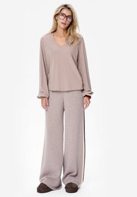Lichtbeige langemouwen top met V-hals; gecombineerd met brede beige broek met zijstrepen. Model draagt bruine schoenen. Gestructureerde stof.