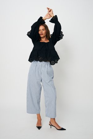 Žena v černé haleně z průsvitné krajky s nabíranými rukávy a bílých širokých kostkovaných kalhotách pózuje s rukama zvednutýma vzhůru.
