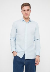 Lys blå langermet skjorte i et glatt stoff med krage som kan kneppes og en enkelt brystlomme, kombinert med mørk blå jeans.