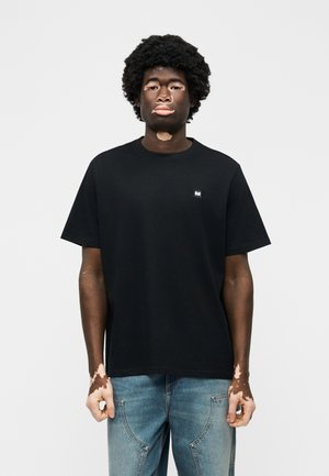 Νεαρός με λεύκη που φοράει ένα απλό μαύρο μπλουζάκι και μπλε τζιν, στέκεται μπροστά σε λευκό φόντο.