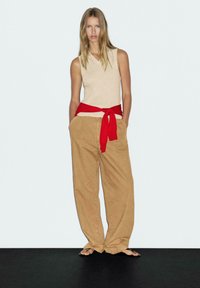 Top beige senza maniche con scollatura a V, cinturino rosso annodato in vita e pantaloni beige a gamba larga. La modella si trova su una superficie scura contro uno sfondo chiaro.