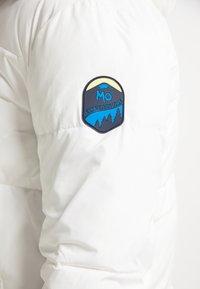 Weißer Pufferjacke mit einem bunten Patch mit Bergen, einer Sonne und dem Text "MO EXPEDITION TIME" auf dem linken Ärmel.