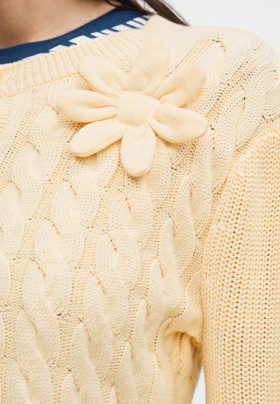 Κρεμ πουλόβερ πλεκτό με σχέδιο καλώδιο και μεγάλο πλεκτό λουλούδι καρφιτσωμένο κοντά στον ώμο πάνω από ένα μπλε πουκάμισο.