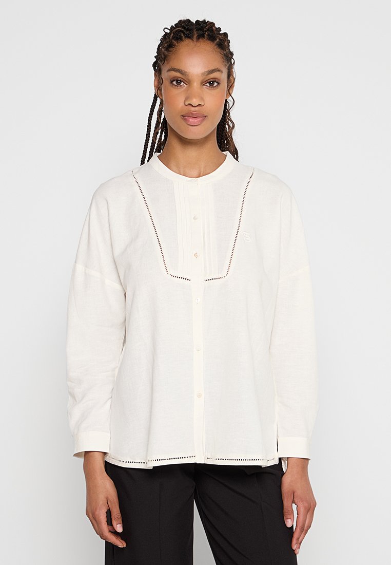 Chemise blanche en lin à boutons avec un col rond, ornée de plis verticaux et de broderies décoratives le long de la patte de boutonnage et de l'ourlet.