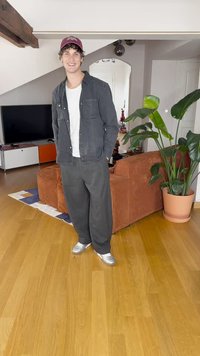 Kjedelig antrekk med en mørk skjorte over en hvit t-skjorte, vide grå bukser og sølvfargede sneakers. Maroon caps og innendørs setting.