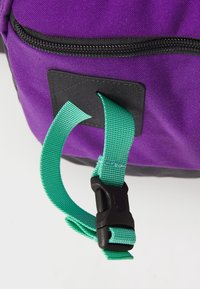 Portion d'un sac à dos violet avec une fermeture éclair noire. Comprend une sangle ajustable verte et un point de fixation noir pour sécuriser des éléments supplémentaires.
