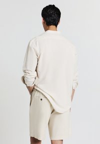 Homme aux cheveux bruns portant une chemise crème à manches longues retroussées et un short crème assorti, debout avec les mains dans les poches.