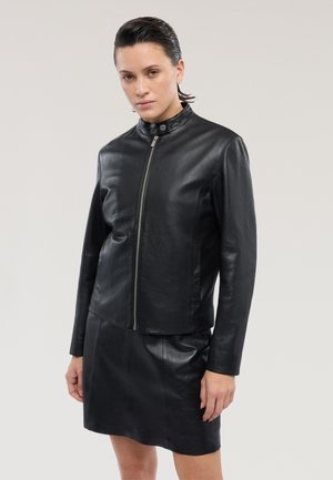 Giacca in pelle nera con collo alto, chiusura con zip argentata e design aderente. Abbinata a una gonna in pelle coordinata per un look armonioso.