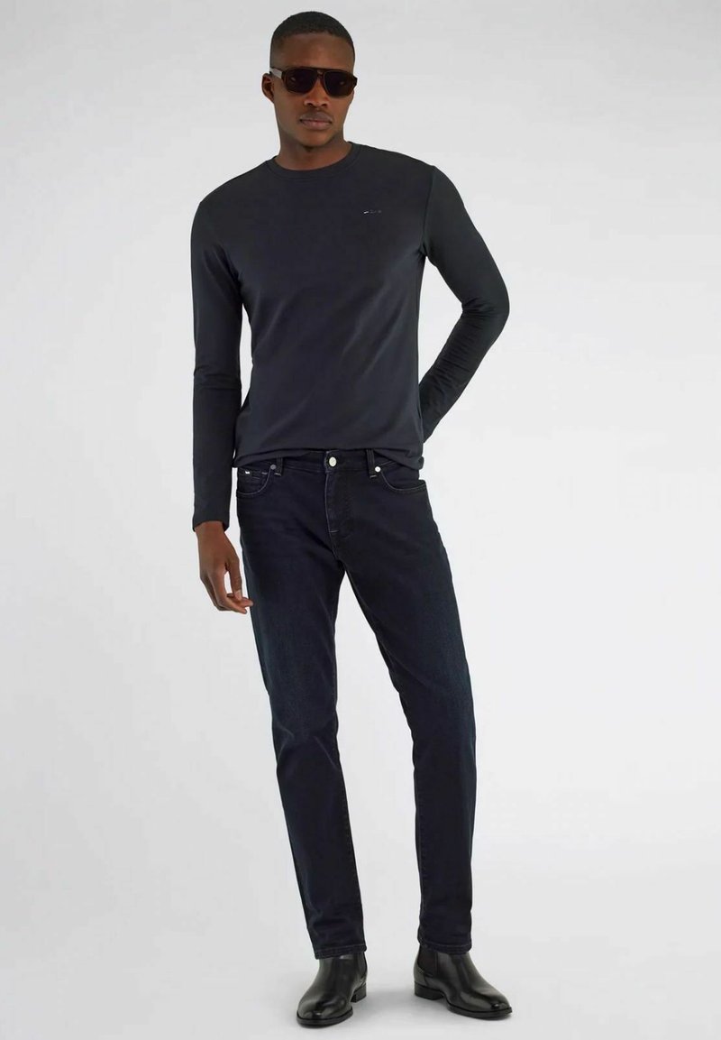 Camicetta nera a maniche lunghe abbinata a jeans scuri. Il modello indossa occhiali da sole e stivaletti neri. Design semplice, senza motivi o loghi visibili.