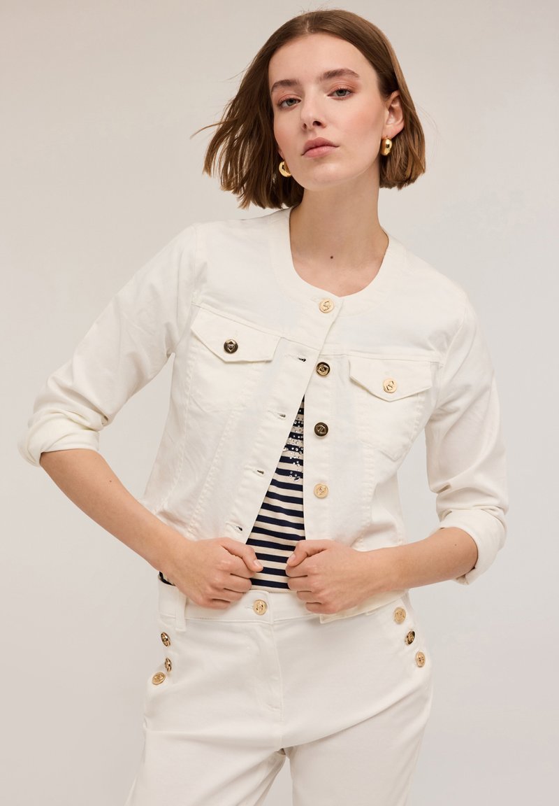 Vit denimjacka med guldknappar och två bröstfickor, bärs över en marinblå och vit randig topp. Modell med kort hår poserar.