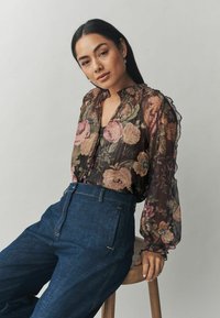 Permatomas juodas gėlių bliuzonas su rožinėmis ir mėlynomis rožėmis, volangų apykakle ir pūstomis rankovėmis, derinamas su aukštu liemeniu tamsiomis džinsinėmis kelnėmis.