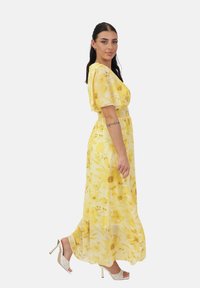 Gul maxikjole med blomsterprint, korte puffede ærmer og smockdetalje ved taljen. Stoffet har en let tekstur og en flydende silhuet. Kombineret med hvide sko med hæl.