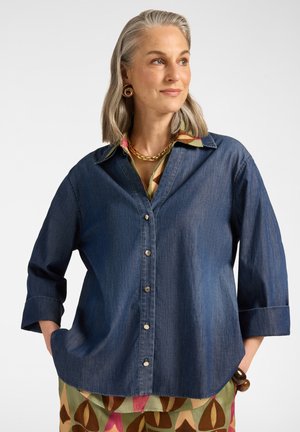 Frau mit grauen Haaren, die ein blaues Jeanshemd über einem bunten, gemusterten Kleid trägt, mit goldener Halskette und großen runden Ohrringen, blickt zur Seite.