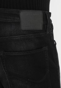 Mustat farkut, joissa on tekstuuroitu pinta, nahkapaneeli takana, takataskut ja hienovaraiset ompeluyksityiskohdat.