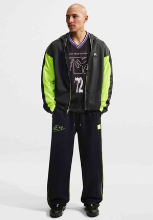 Mand med barberet hoved iført en sort og neongrøn jakke, en sort grafisk t-shirt, sorte bukser med neon detaljer og sorte sneakers.