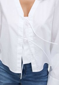 Camicia bianca in cotone con scollatura a V profonda, dettaglio arricciato e legato in vita, e maniche lunghe, abbinata a jeans blu in denim.