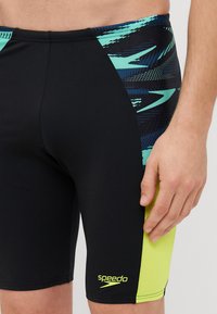 Speedo peldēšanas šorti melnā krāsā ar zilu un aqua rakstu sānu paneļos, kam ir spilgti dzeltens akcents un logo apakšējā labajā stūrī.
