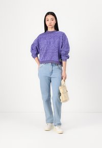 Lila sweatshirt med ett strukturerat geometriskt mönster, ljusblå jeans och en liten beige väska. Vita sneakers fulländar outfiten.