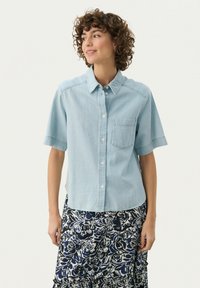 Light blue short-sleeve denim shirt with a button-down collar, chest pocket, and subtle stitching details. Worn with a patterned floral skirt.