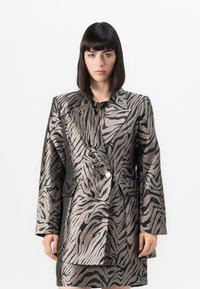 Kvinne med rett, mørkt hår iført en metallisk zebra-mønstret blazer og matchende skjørt, stående mot en hvit bakgrunn.