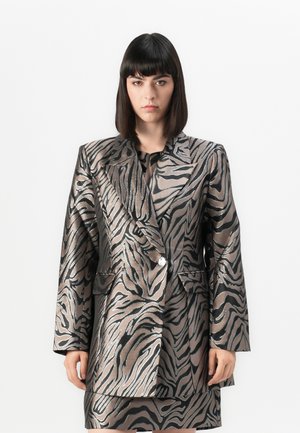 Woman with straight dark hair wearing a metallic zebra-print blazer and matching skirt, standing against a plain white background.
