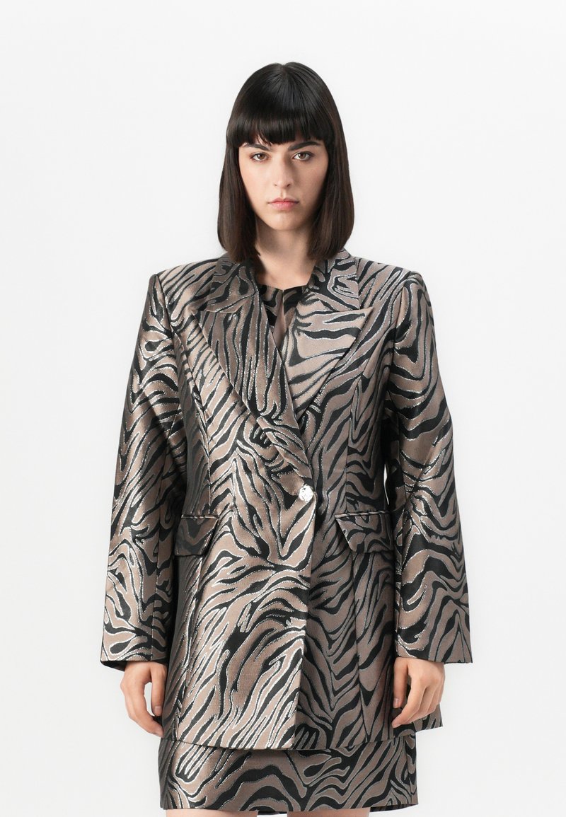 Kvinne med rett, mørkt hår iført en metallisk zebra-mønstret blazer og matchende skjørt, stående mot en hvit bakgrunn.